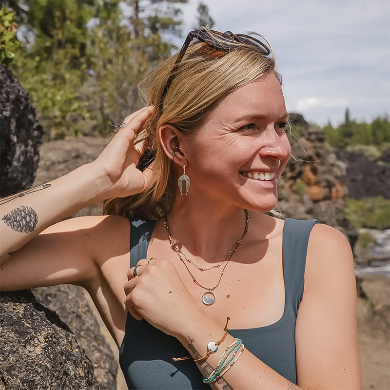 A smiling woman with tattoos in a blue top showcases her waterfall-inspired earrings.