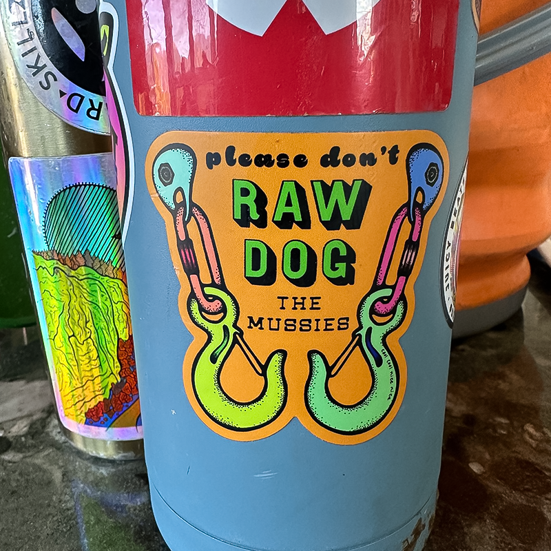 A blue tumbler with an orange bold sticker that says "please don't RAW DOG THE MUSSIES".