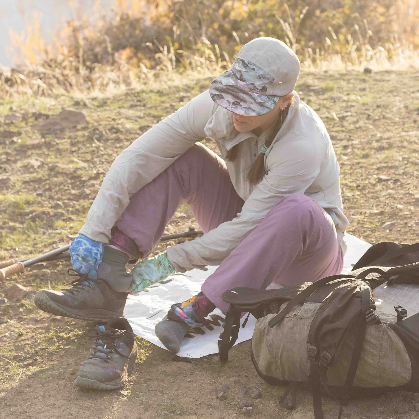 A woman on the ground with a backpack sitting on the Cowboy Camping Dust Buster sit pad while putting on her hiking shoes.