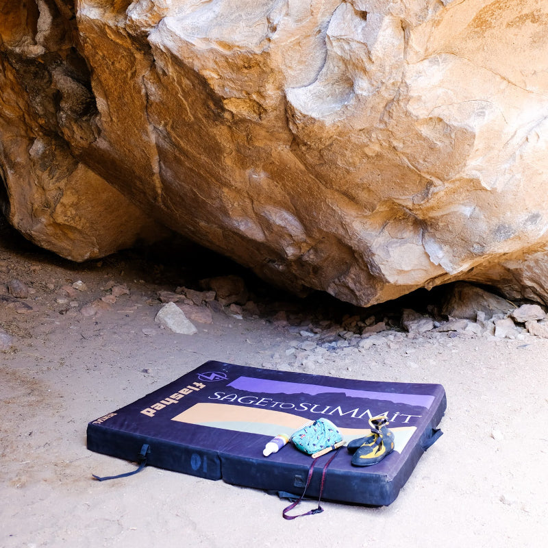 A crash pad with the Sage to Summit logo laying below a boulder with a chalk bag and climbing shoes sitting on it.