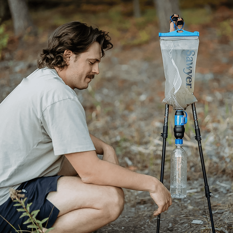A man beside a tripod and water bottle, illustrating the Sawyer Squeeze Water Filtration System.