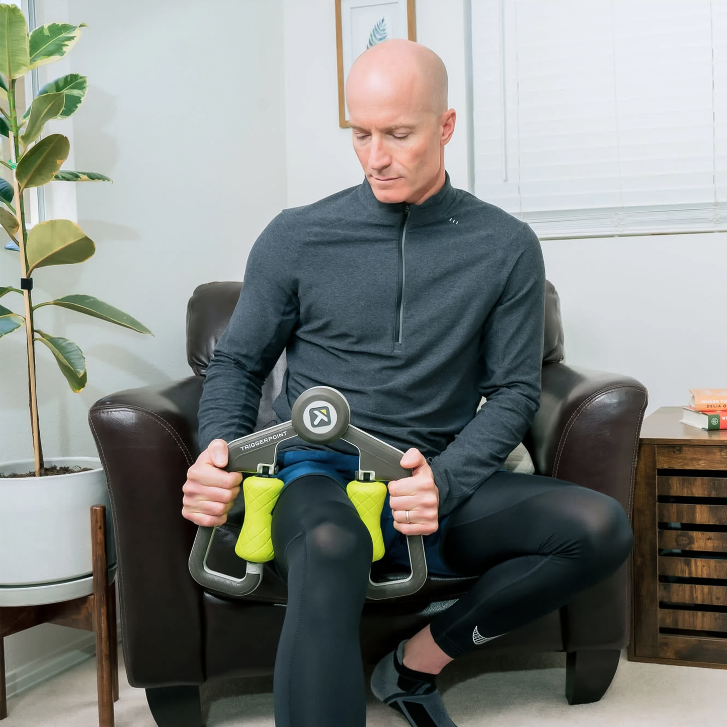 A man in a knee brace sits on a chair, holding the TriggerPoint FUSE Massager for muscle recovery and relief.