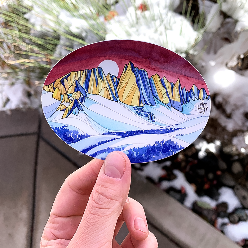 A hand holding a 4x3" oval sticker depicting a mountain scene, designed by Alex Bailey, printed on durable vinyl.