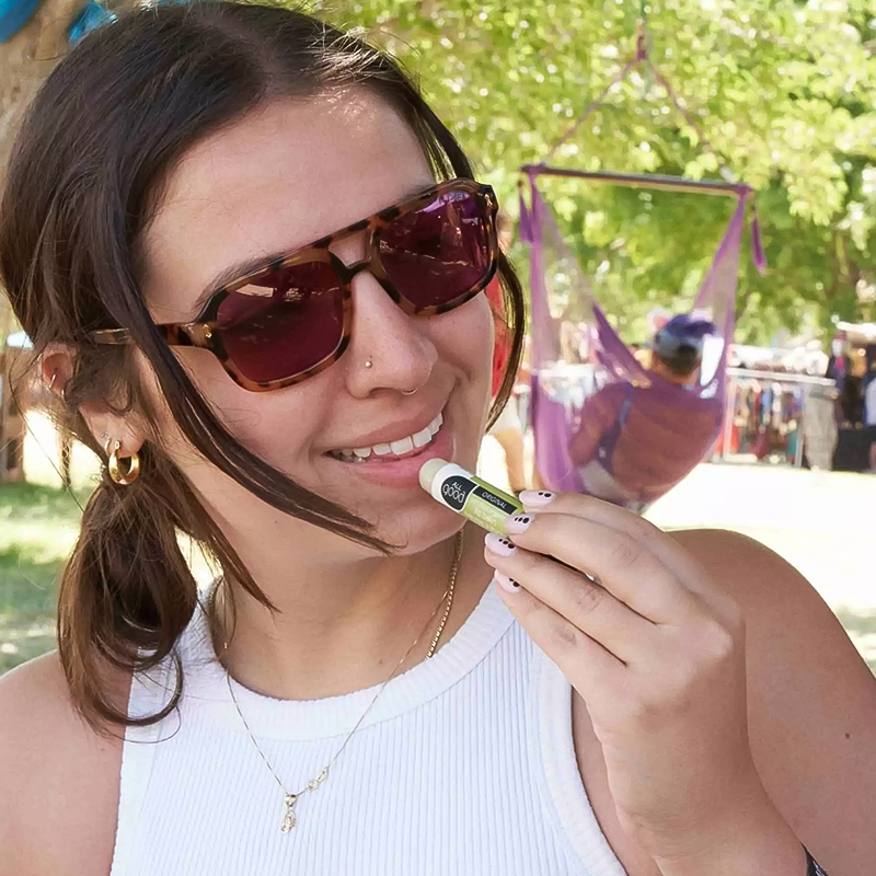 A woman in sunglasses holds an All Good organic original Lips SPF15, a natural lip balm for sun protection.