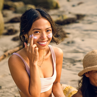 A woman at the beach, smiling and applying Badger Adventure SPF 50 Mineral Sunscreen for sun protection during her outdoor fun.