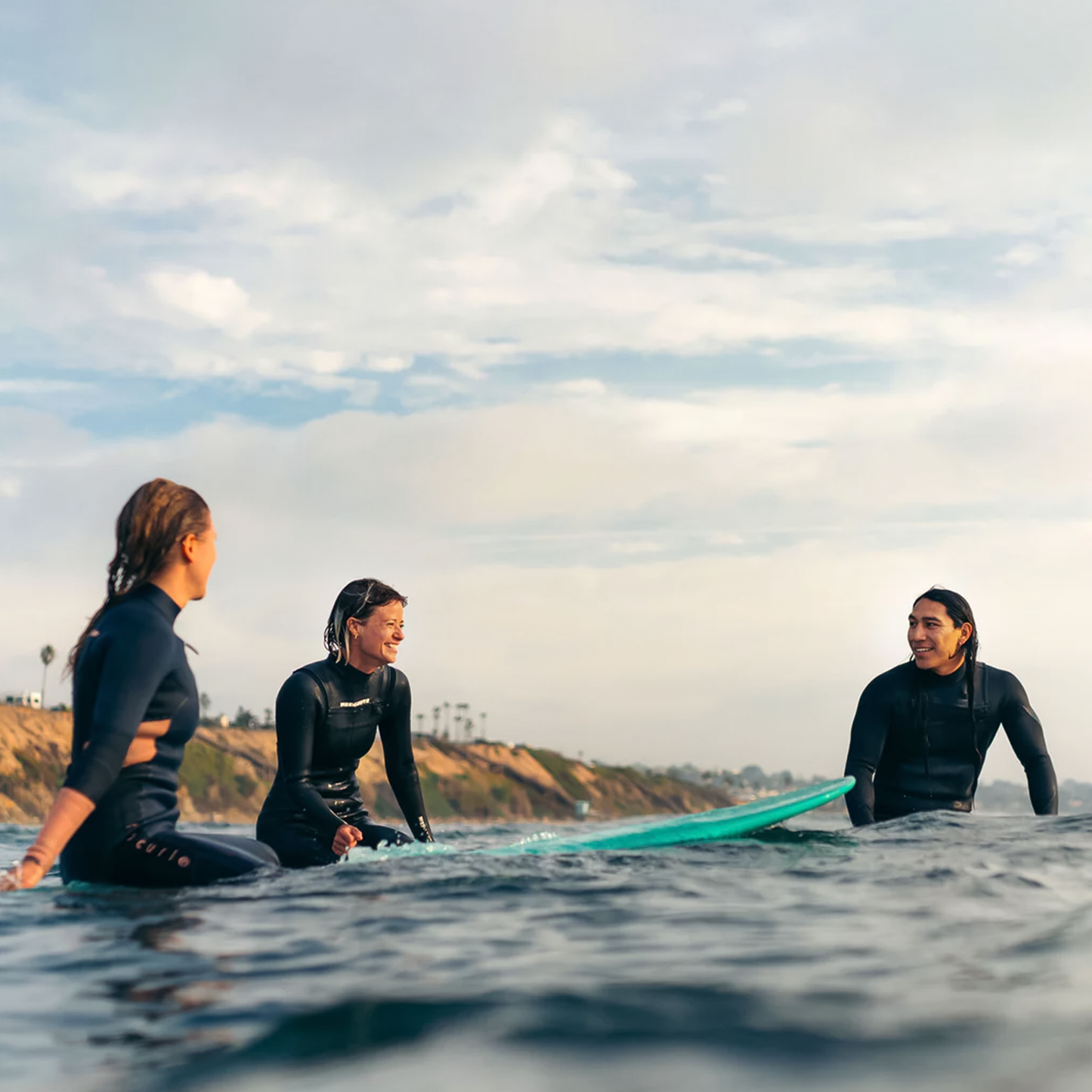 Three people in wetsuits sit on surfboards, smiling and enjoying a sunny day at the beach, ready for surfing adventures.