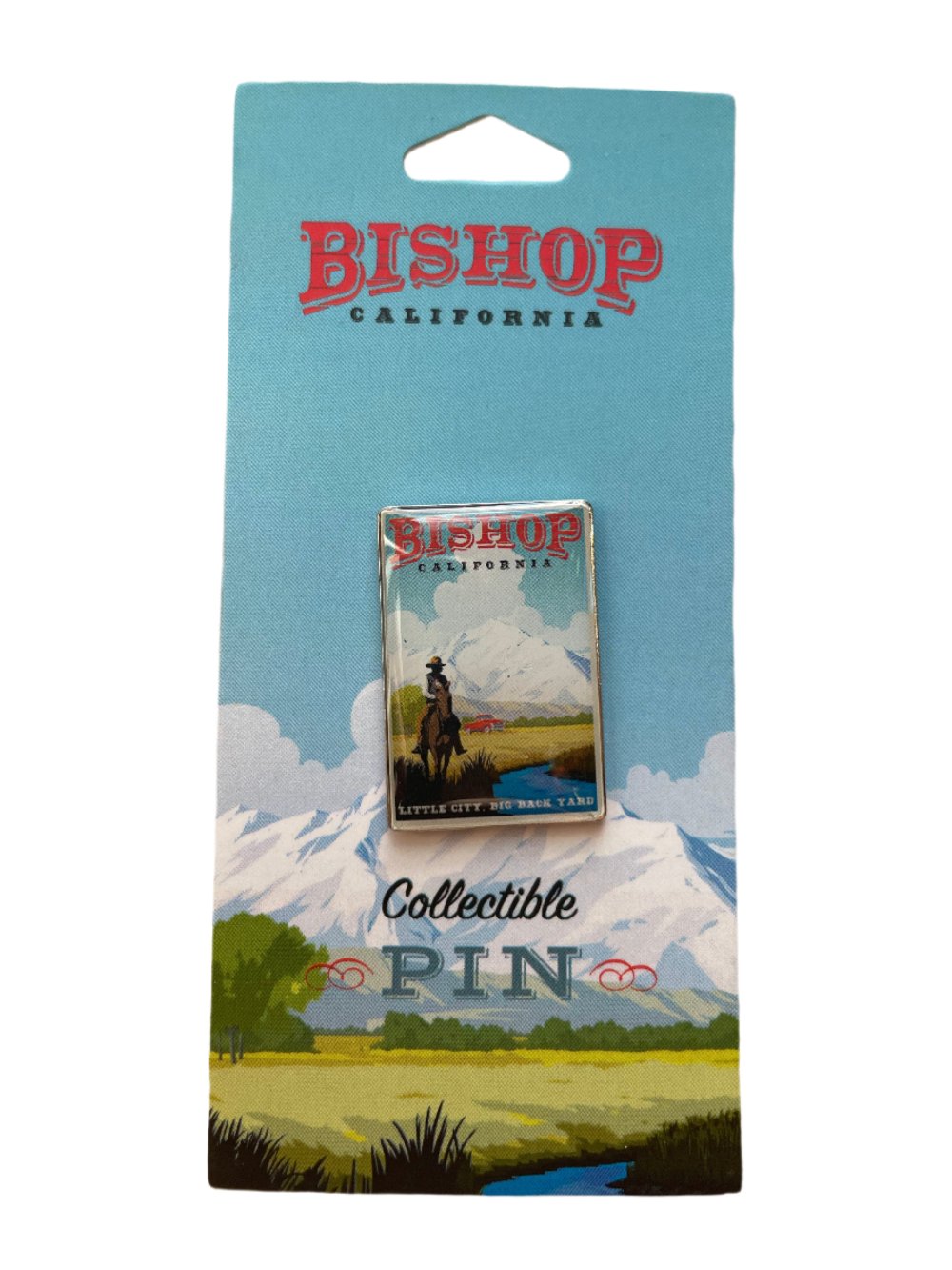 A collectible metal pin for Bishop, California depicting a horse and rider next to a river in front of a mountain scene. The bottom says &quot;little city, big backyard.&quot;