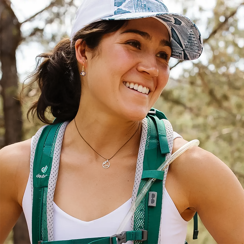 A smiling woman in a hat and backpack showcasing a black nylon necklace with sterling silver mountain-inspired pendant, embracing nature's beauty.