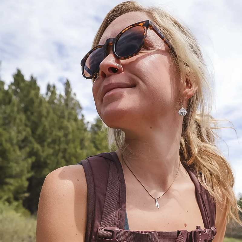 A woman in sunglasses and a backpack, showcasing her adventurous spirit and style with mountain-themed earrings.
