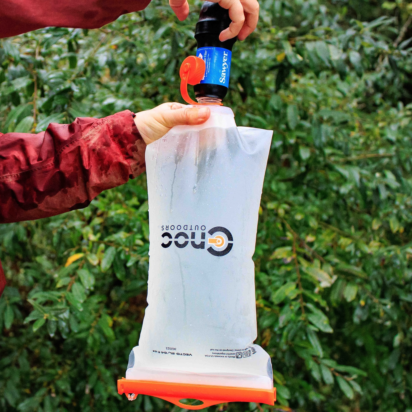 A person in an red jacket holds a clear and orange CNOC 28mm Vecto 2L Water Container by CNOC, with a water filter attached on top, filling it outdoors amid green foliage.