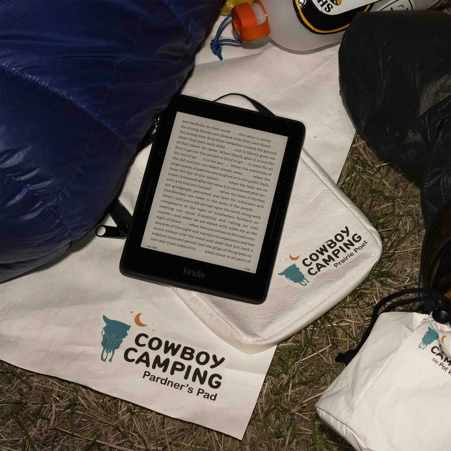 A Kindle e-reader and a puch are placed on the ground beside a sleeping bag, showcasing a travel-friendly setup.