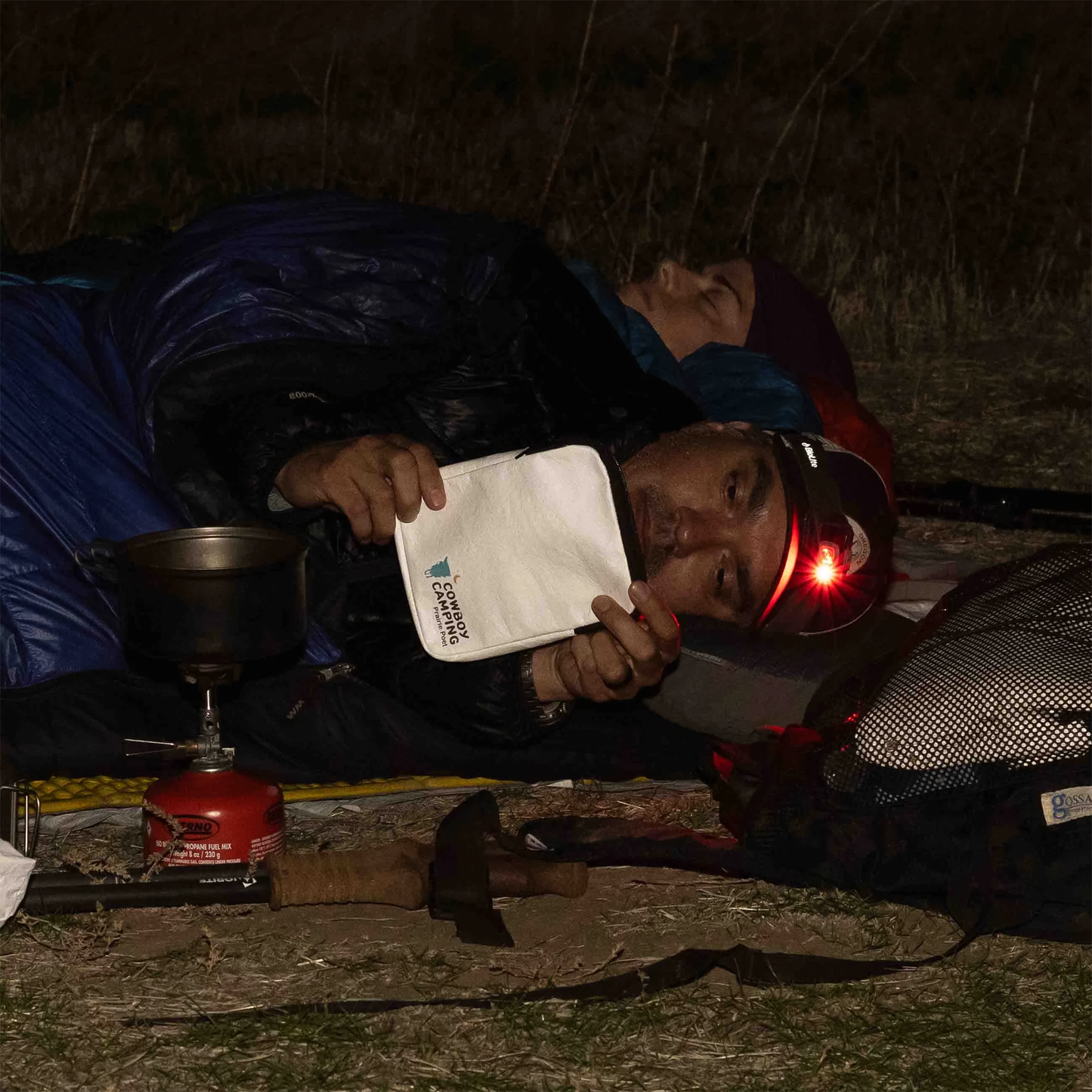 A man reads on an e-reader while holding a pouch beside a woman sleeping on a sleeping bag outdoors, creating a peaceful camping scene.