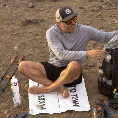 A man sits on the Cowboy Camping Dust Buster sit pad on the ground with a backpack for comfort during a break outdoors.