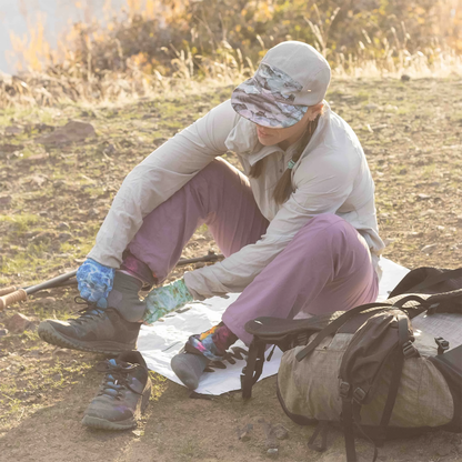 A woman on the ground with a backpack sitting on the Cowboy Camping Dust Buster sit pad while putting on her hiking shoes.