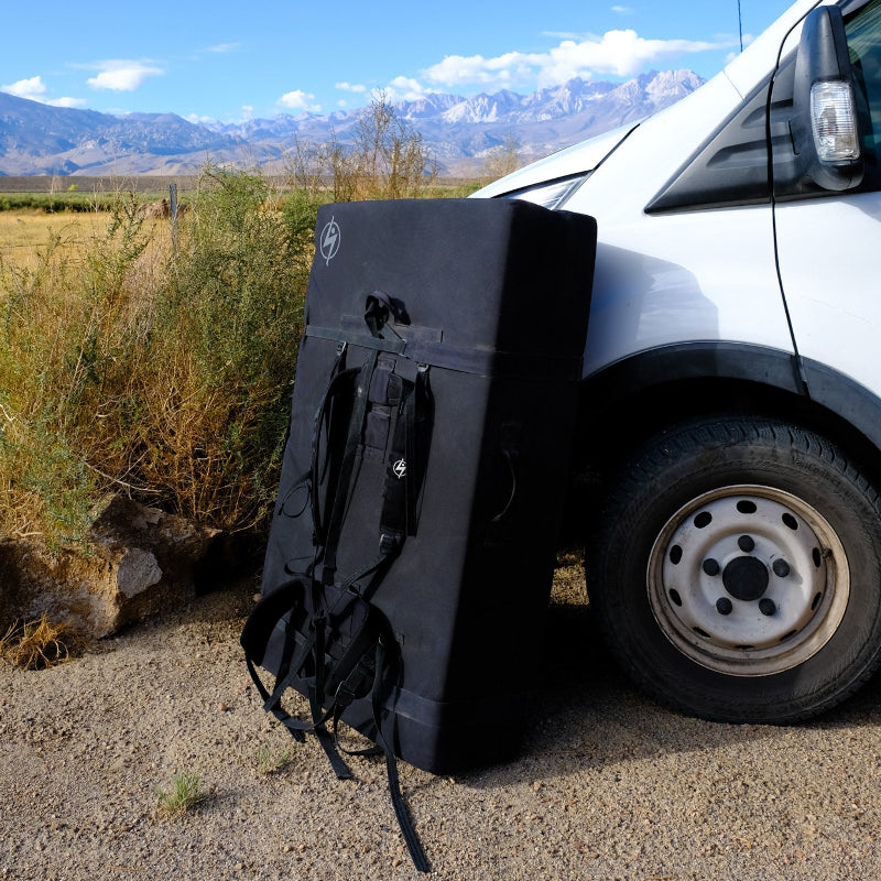 A closed bouldering crash pad leaning against a car with mountains in the background.