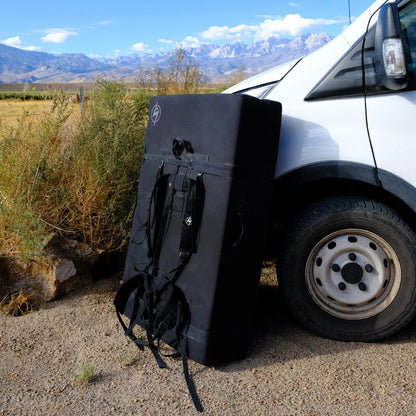 A closed bouldering crash pad leaning against a car with mountains in the background.