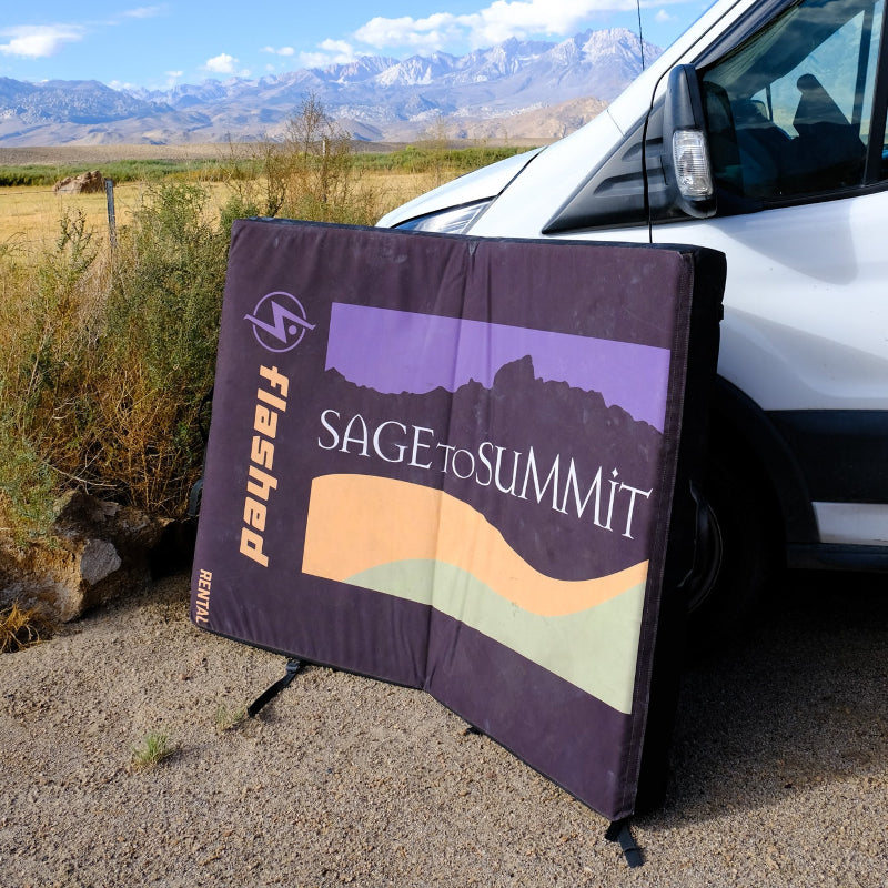 Bouldering pad with the Sage to Summit logo laying open against a car with mountains in the background.