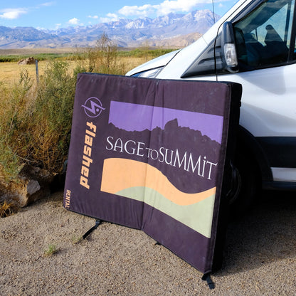 Bouldering pad with the Sage to Summit logo laying open against a car with mountains in the background.