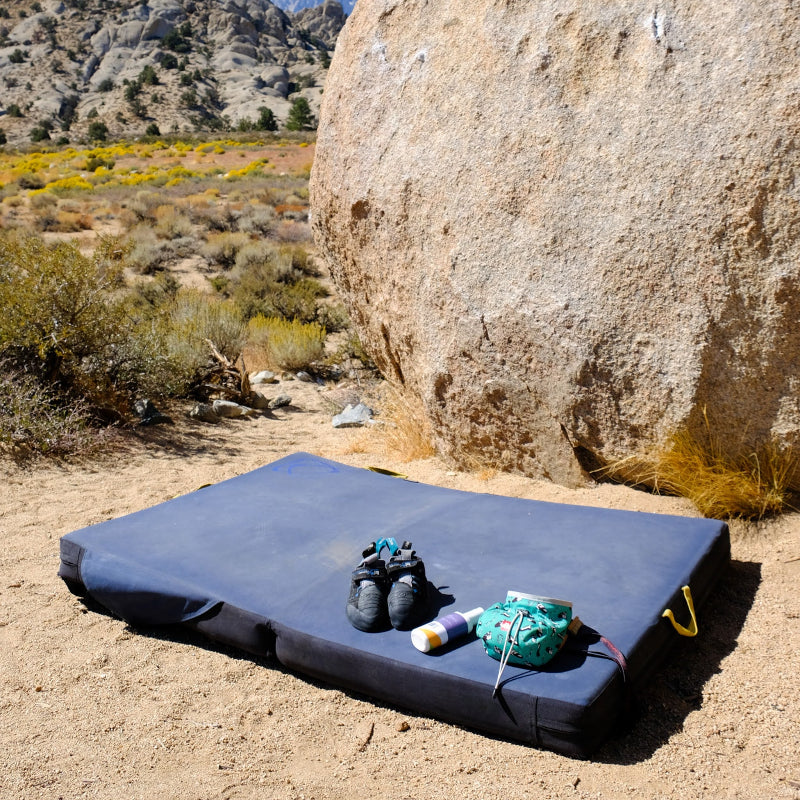 A large blue crash pad for bouldering laying below a boulder with climbing shoes and a chalk bag laying on it.