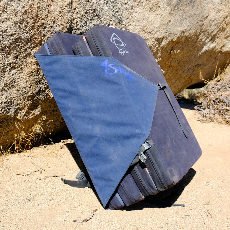 A smaller crash pad strapped to a large crash pad, both leaning against a boulder. 