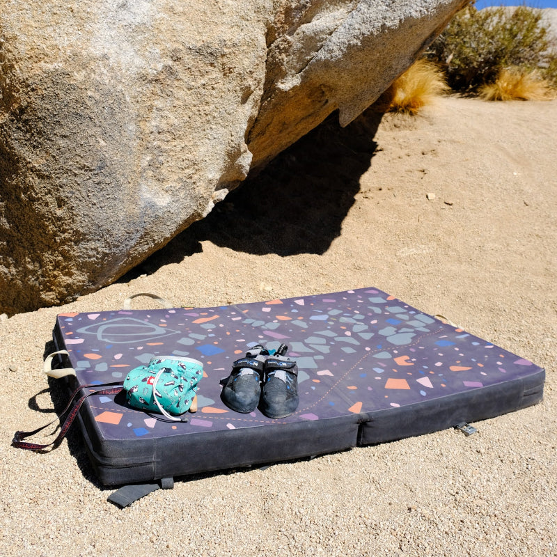 A multicolored Asana bouldering pad laying below a large boulder with a chalk bag and climbing shoes sitting on it.