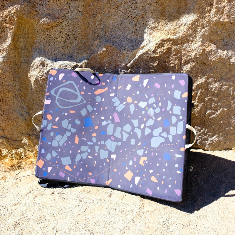 A speckled Asana bouldering pad laying open against a large boulder. 