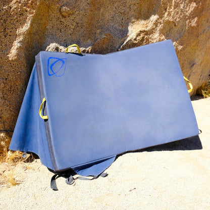 A large blue Asana crash pad for climbing leaning against a rock wall.