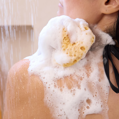 A woman lathers her back with Dr. Bronner’s Peppermint Liquid Soap in a bright bathroom setting.