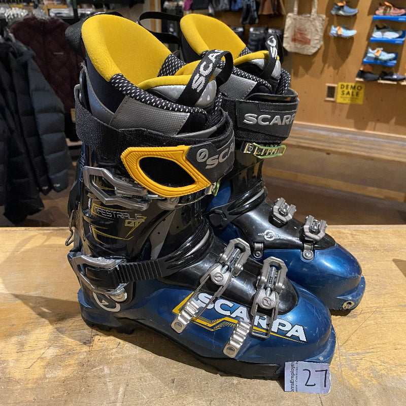 Pair of blue and black ski boots with yellow accents on a wooden floor.