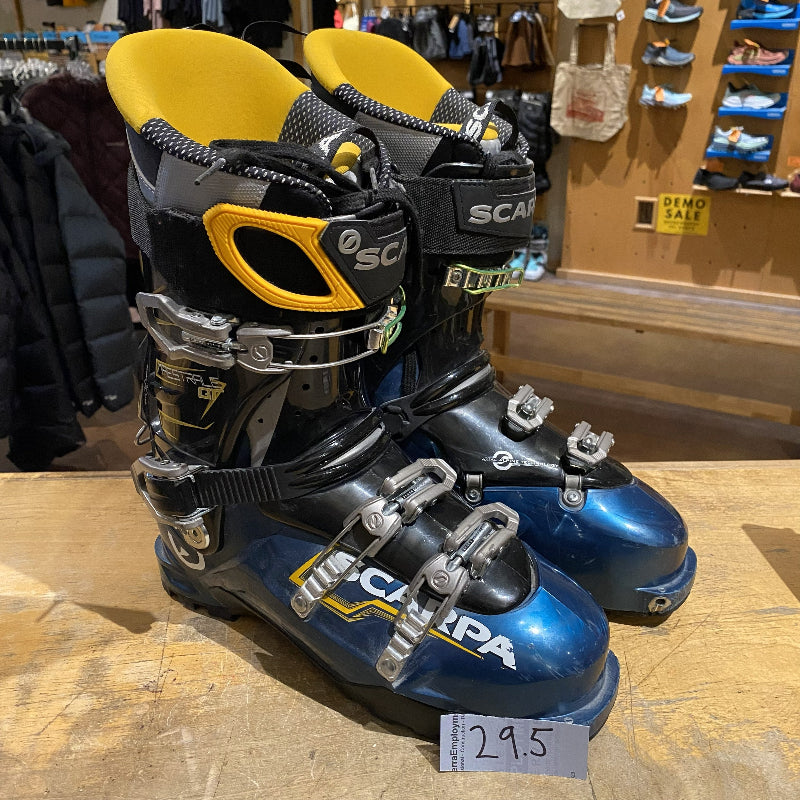 Pair of blue and black ski boots with yellow accents on a wooden floor in a store setting.