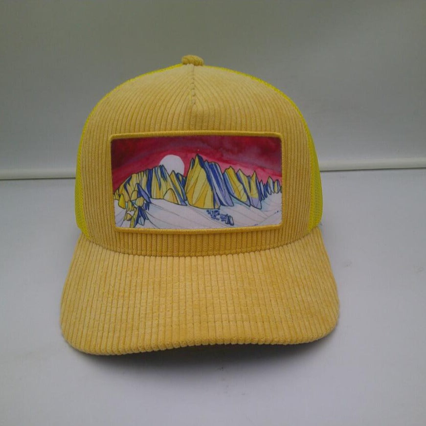 The image showcases the Mammoth Skyline Corduroy Trucker Hat, featuring its khaki color top and undervisor, gold front panel, navy undervisor, and the unique patch design of the Minerats of Mammoth Lakes by Alex Bailey Art.