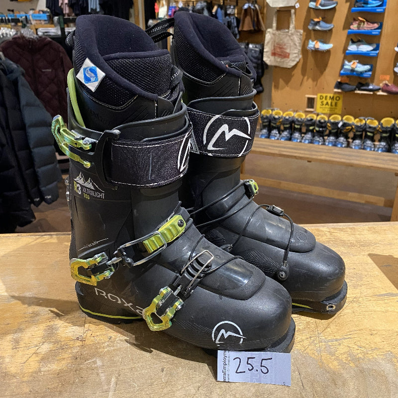 Pair of black ski boots with green straps on a wooden surface in a store setting.