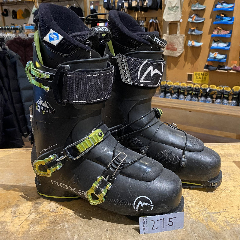 Pair of black ski boots with green buckles on a wooden surface in a store setting.