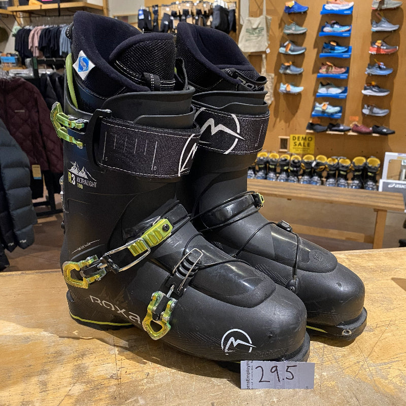 Pair of black ski boots with yellow accents on a wooden floor in a store setting.