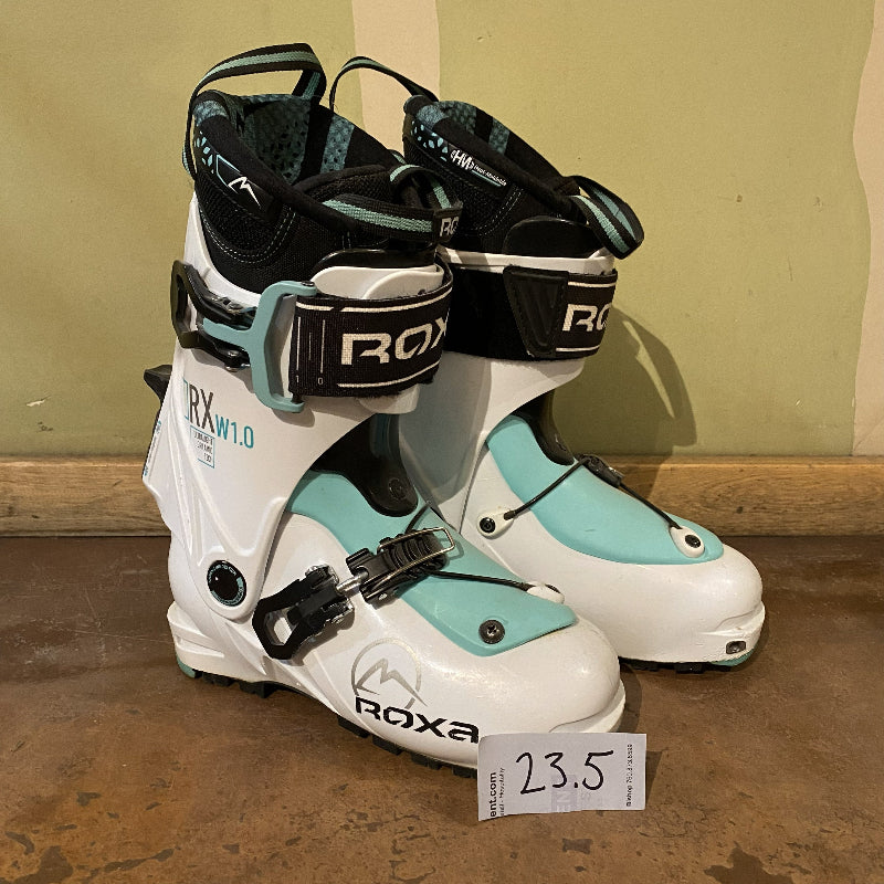 A pair of white backcountry ski boots against a green wall. 