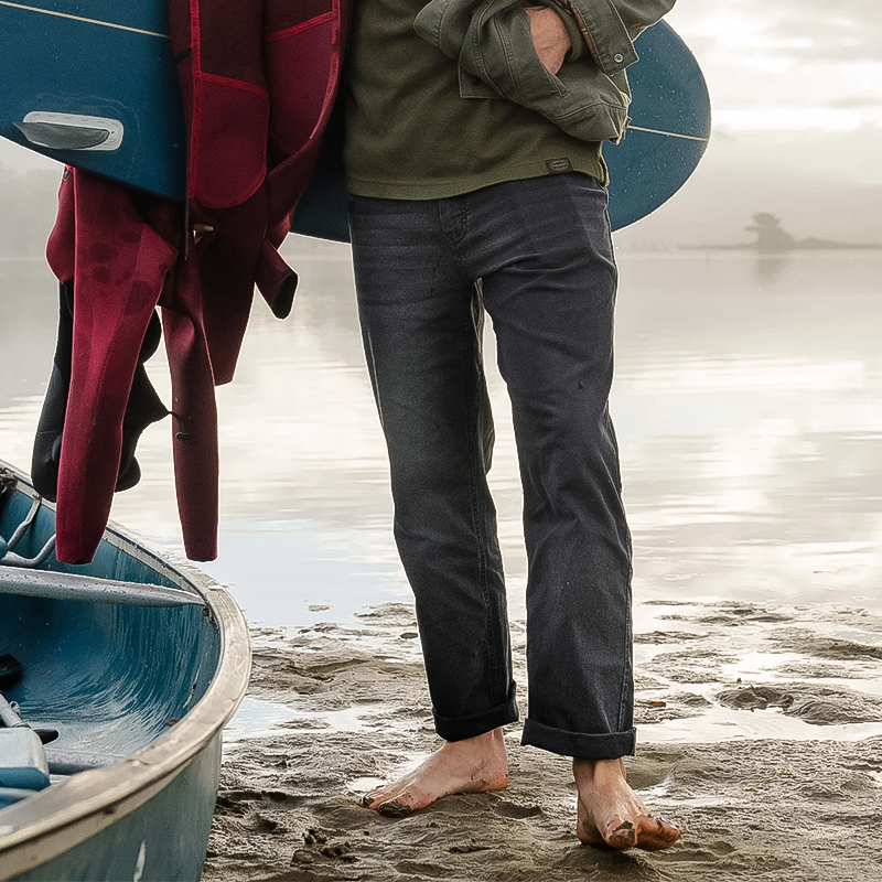 A man beside a boat holds a surfboard, wearing relaxed-fit Ripton Superlite Jeans.
