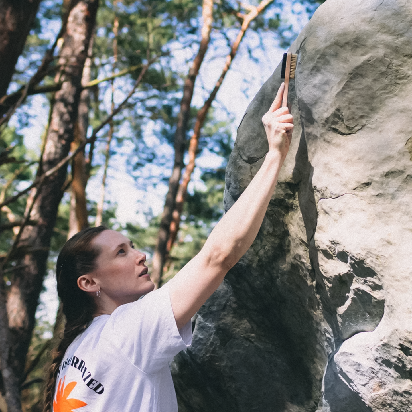 A woman standing beside the rock in the woods while using the Rúngne Double Boaring Chalk Brush for cleaning holds.