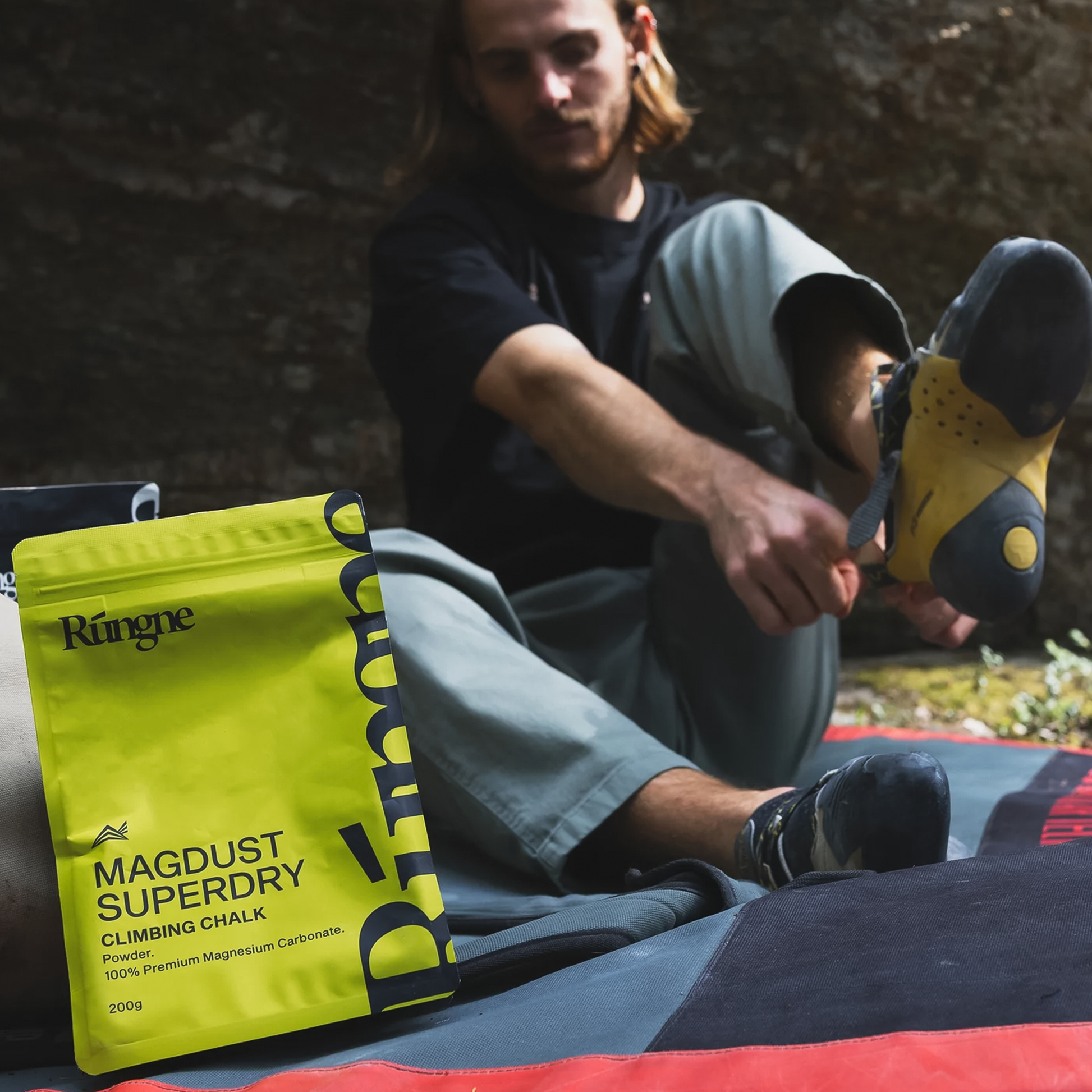 A man sits on a blanket while putting on a climbing shoe with a pack of Rungne Magdust Superdry Chalk for climbing.