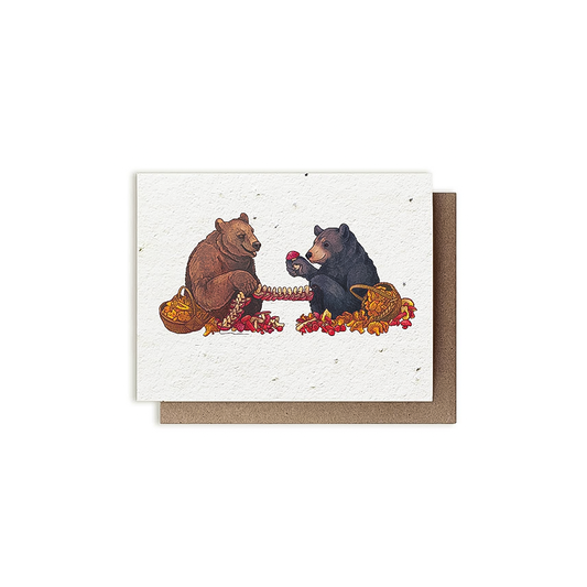 A brown and black bear illustrated on a card enjoy apples, surrounded by mushrooms and plantable paper.