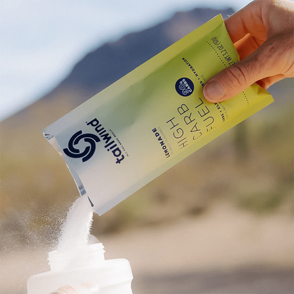A hand holds Tailwind High Carb Fuel lemonade powder in front of a mountain, ready for training.  