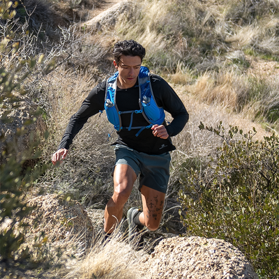 [Image Description: The Ultimate Direction Mountain Vest 6.0 is shown in action on a trail runner navigating a scenic mountain path. The vest fits snugly around their torso and showcases its various pockets and compartments for easy access to essentials.]