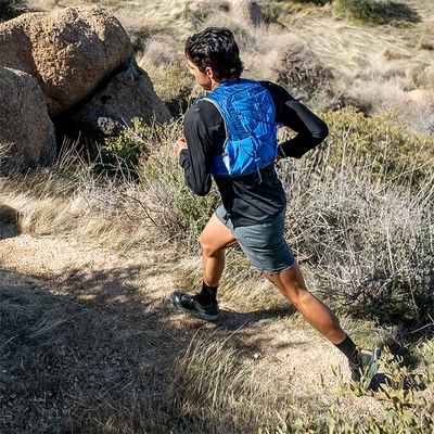 [Image Description: The Ultimate Direction Mountain Vest 6.0 is shown in action on a trail runner navigating a scenic mountain path. The vest fits snugly around their torso and showcases its various pockets and compartments for easy access to essentials.]